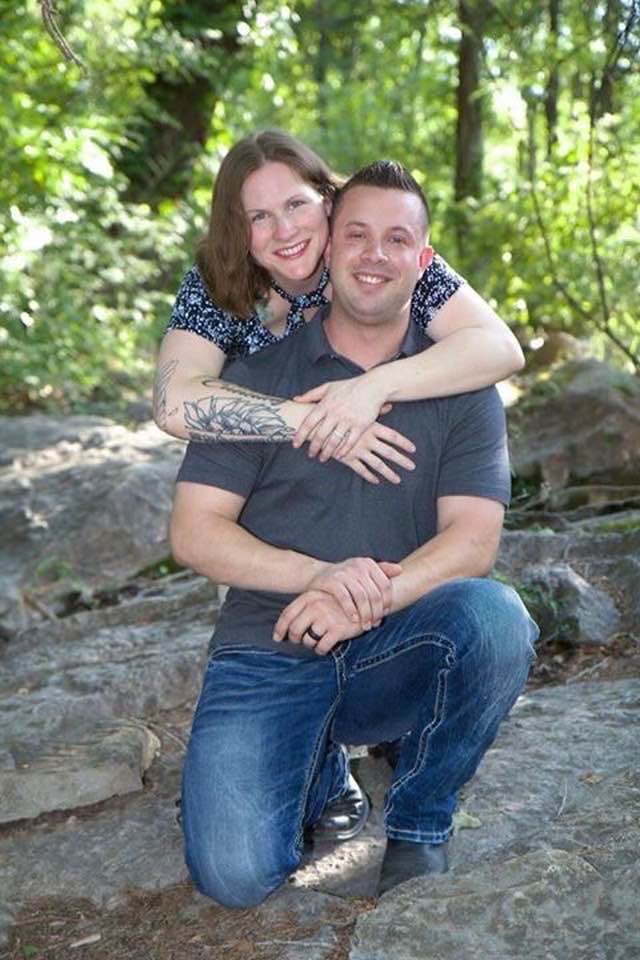 Smiling couple embracing outdoors on a rocky path surrounded by green forest trees.